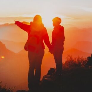 An image of a mother and 7 year old son holding hands, the mother is pointing out to the mountains, the sun is setting and the sunset color is orange