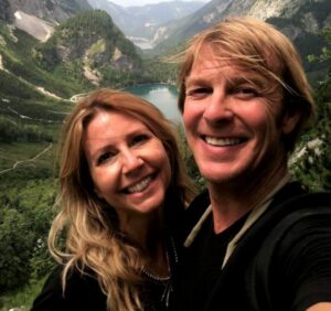 Blonde hair woman and blonde hair man in black t-shirts happy, smiling, and hiking in the alps in Austria near a lake, this image is of the owners of Certified Travel International